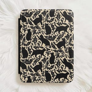 May include: A rectangular e-reader case featuring a repeating pattern of black cat silhouettes interspersed with black leafy foliage on a cream-colored background. The cover has a textured surface and rounded corners, with a black spine visible on the left.