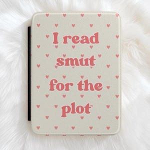 May include: A rectangular e-reader case with a textured cream-colored surface featuring a repeating pattern of small pink hearts. The text "I read smut for the plot" is printed in a bold, pink, rounded font across the center of the front cover.