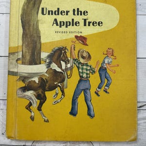 May include: A hardcover book titled Under the Apple Tree Revised Edition with a yellow cover featuring an illustration of a brown and white horse, a person in a plaid shirt and cowboy hat holding a rope, and another person running nearby.