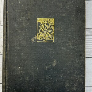May include: A black hardcover book with a gold-colored illustration on the front cover. The illustration depicts a person playing a flute in a garden setting. The text "TOVT BIEN OV BIEN" is visible on the illustration.