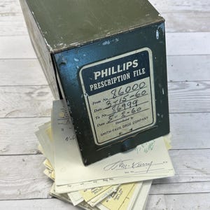 May include: An olive green metal Phillips Prescription File box with a label showing dates and numbers. The box is slightly open, revealing a stack of old prescription papers. The box is labeled "SMITH-FAUS DRUG COMPANY."