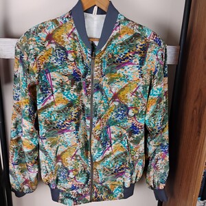 May include: A patterned bomber jacket in a mix of teal, yellow, purple, and blue. The jacket features a grey collar, cuffs, and hem, and a full-length zip. It is hanging on a wooden hanger. The jacket has an abstract design.