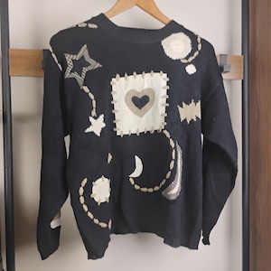 May include: Black sweater with cream and tan celestial patches, including a heart, stars, and moon. The patches are stitched onto the sweater, giving it a handcrafted appearance. The sweater is displayed on a wooden hanger, showcasing its design.