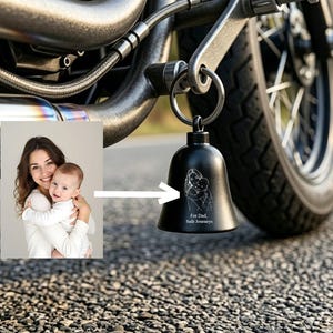 May include: Black motorcycle bell with a family illustration and the text "For Dad, Safe Journeys." The bell is attached to a motorcycle. A photo of a mother and child is in the background.