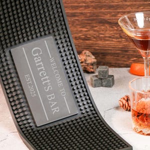 May include: A black bar mat with a textured surface and the text "WELCOME TO Garrett's BAR EST.2025". A cocktail and a glass of whiskey are on the table. Also, there are ice cubes and a slice of fruit.