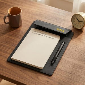 May include: A black leather clipboard with a notepad and pen, featuring the initials "WIM" in gold. The notepad has the text "Aesthetic Observations: Initial thoughts for the new collection..." A gold-colored analog clock and a brown mug are also on the wooden surface.