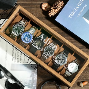 Peut inclure: Un coffret en bois pour montres, de 31 cm de long et 12 cm de large, avec un dessus en verre, contenant plusieurs montres-bracelets. L'intérieur est marron clair et le texte "Adien Moments That Do Us" est visible.