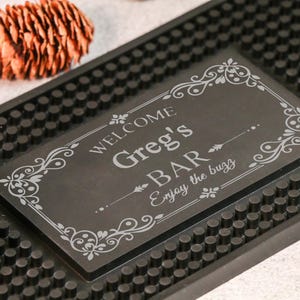Può includere: Un tappetino da bar nero con la scritta "WELCOME Greg's BAR Enjoy the buzz" in un carattere decorativo. Il tappetino ha una superficie strutturata e un bordo decorativo. Una pigna è sullo sfondo.