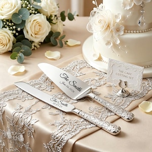 May include: A silver cake knife and server set with engraved text. The set includes a serrated knife and a wide server, both with ornate handles. The text on the server reads "The Smith June 06 2026". The knife reads "The Smith 10.03.25".