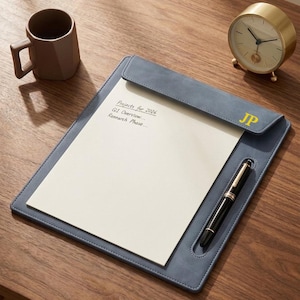 Könnte beinhalten: Ein blaues Leder-Schreibtischunterlage mit einem weißen Notizblock, einem schwarzen Stift und den Initialen "JP" in Gold. Der Notizblock hat handschriftlichen Text mit der Aufschrift "Projects for 2026, Q1 Overview, Research Phase...". Eine goldene Uhr und eine braune Tasse befinden sich ebenfalls auf dem Holzschreibtisch.
