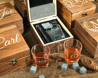 May include: Wooden gift boxes with personalized names and initials are displayed. One box is open, revealing a whiskey glass, ice cubes, and tongs. Two glasses of amber liquid sit in front, with ice cubes and pine cones.