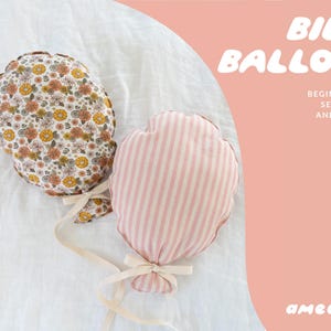 Puede incluir: Dos peluches hechos a mano con forma de globo. Uno tiene un diseño floral en tonos marrones, amarillos y blancos. El otro es de rayas rosas y blancas. Ambos tienen "cuerdas" de cinta color crema y están atados con lazos. El texto dice "Billie Balloon".