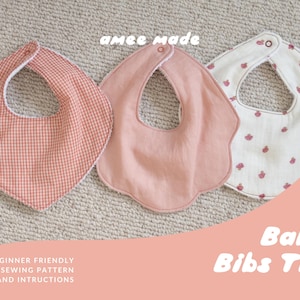 May include: A trio of baby bibs featuring a coral gingham check bib, a solid dusty rose scalloped bib, and a white bib with a small red fruit print. Each bib includes a snap closure at the neck. The text "amee made" and "Baby Bibs Trio" is visible.