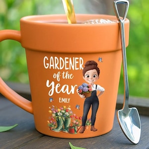 May include: An orange ceramic mug shaped like a flower pot with a handle, featuring the text GARDENER of the Year EMILY, an illustration of a person holding a basket of flowers, and a silver shovel-shaped spoon resting against the side.