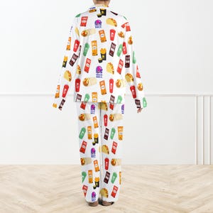 May include: White pyjama set with a repeating pattern of Taco Bell food items and logos. The long-sleeved top and trousers display colourful illustrations of tacos, burritos, fries, and drink cups. The pyjama set is designed for comfort and casual wear.