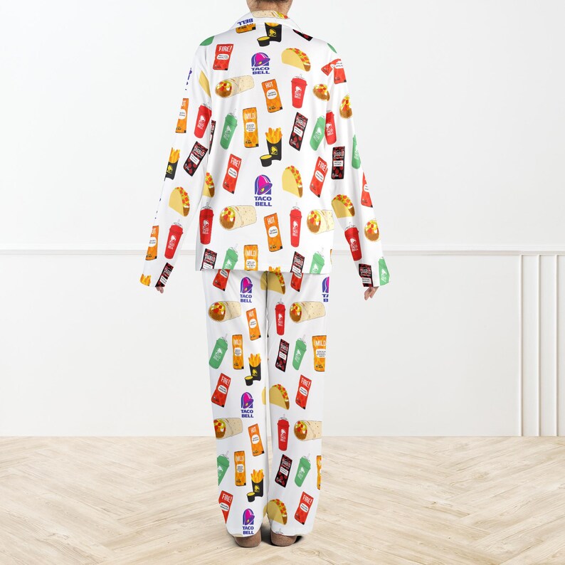 May include: White pyjama set with a repeating pattern of Taco Bell food items and logos. The long-sleeved top and trousers display colourful illustrations of tacos, burritos, fries, and drink cups. The pyjama set is designed for comfort and casual wear.