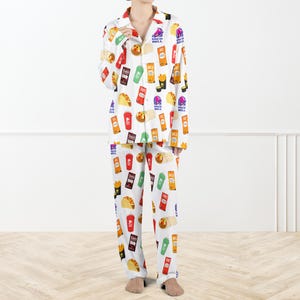 May include: White pyjama set featuring a repeating pattern of Taco Bell food items. The long-sleeved top has a collared neckline and button-down front. The trousers are full-length. The pattern includes tacos, fries, and drink cups.