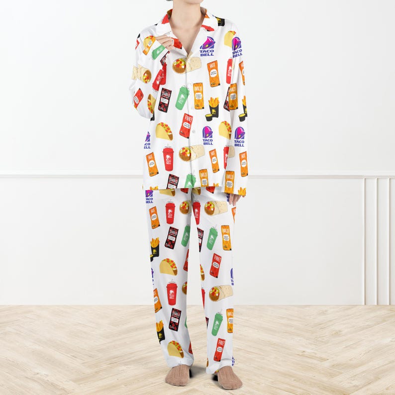 May include: White pyjama set featuring a repeating pattern of Taco Bell food items. The long-sleeved top has a collared neckline and button-down front. The trousers are full-length. The pattern includes tacos, fries, and drink cups.