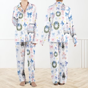 Puede incluir: Conjunto de pijama blanco con estampado festivo navideño. La parte superior de manga larga y los pantalones presentan un estampado de lazos azules, coronas, hombres de jengibre y cascanueces. El pijama tiene una parte superior abotonada y un corte holgado.