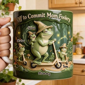 May include: Green ceramic mug with a frog design. The mug features a large frog riding a scooter, with smaller frog characters and the text "Off to Commit Mom Foolery." Names "Chloe," "Mateo," and "Tom" are also visible.