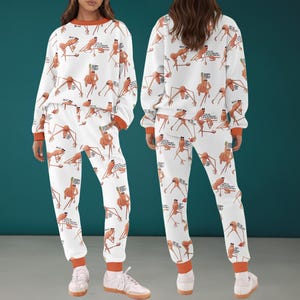 以下が含まれることがあります： 「Sorry, Stuck」のテキストが入った、棒人間の漫画イラストが繰り返される白いパジャマセット。長袖トップスとパンツは、袖口と襟にオレンジ色のトリムが付いています。パジャマセットは人が着用しています。