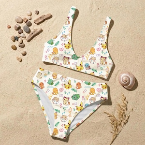 Puede incluir: Conjunto de bikini de dos piezas con un patrón repetido de personajes de dibujos animados coloridos sobre un fondo blanco. La parte superior es de estilo cuello redondo y la parte inferior es de corte clásico. El bikini se muestra sobre arena.