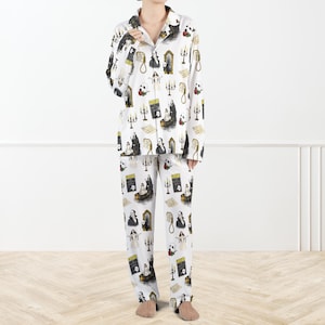 May include: White pyjama set featuring a repeating pattern of gothic illustrations, including portraits, candles, and a panda with a skull. The long-sleeved top has a button-down front and a notched collar. The trousers have a relaxed fit.