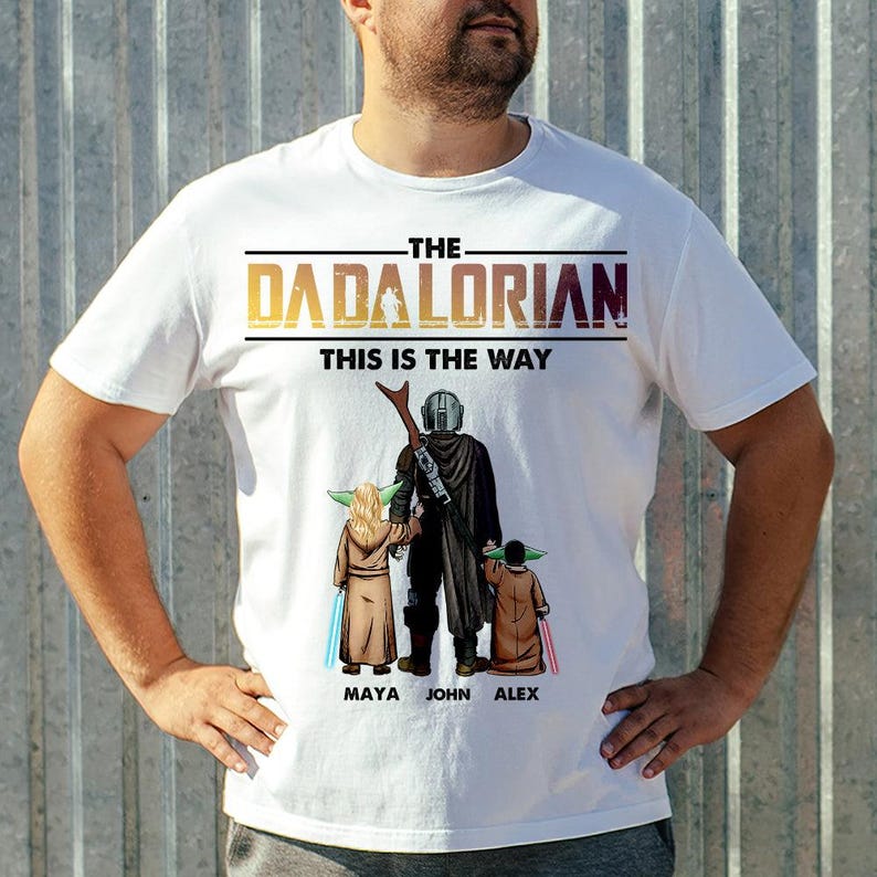 May include: White t-shirt featuring a graphic of The Mandalorian with two Grogu characters and the text "THE DADALORIAN THIS IS THE WAY". The design includes the names Maya, John, and Alex below the characters.