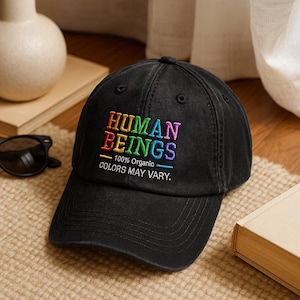 May include: Black baseball cap with the embroidered text "HUMAN BEINGS" in rainbow colors. Below the text are the words "100% Organic" and "COLORS MAY VARY."
