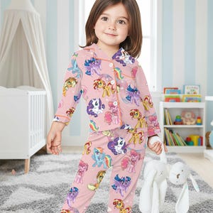 May include: Pink pyjama set featuring cartoon ponies in various colours. The long-sleeved top and trousers are decorated with pony characters. The pyjamas have orange trim and are worn by a young child holding a white stuffed bunny.