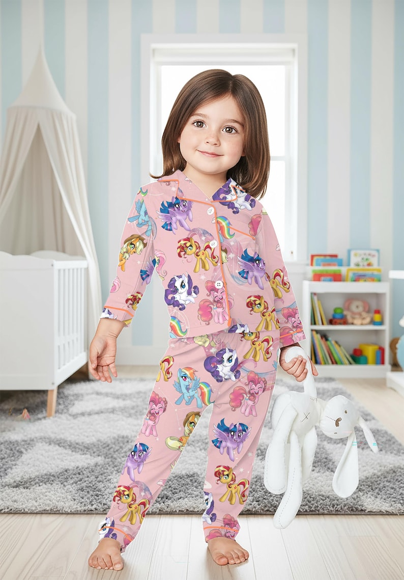 May include: Pink pyjama set featuring cartoon ponies in various colours. The long-sleeved top and trousers are decorated with pony characters. The pyjamas have orange trim and are worn by a young child holding a white stuffed bunny.