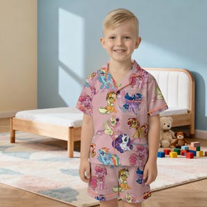 May include: A pink pajama set with a pattern of cartoon ponies. The short-sleeved top and shorts have an all-over print of various pony characters. The set has orange trim and is worn by a young child.