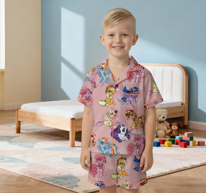 May include: A pink pajama set with a pattern of cartoon ponies. The short-sleeved top and shorts have an all-over print of various pony characters. The set has orange trim and is worn by a young child.