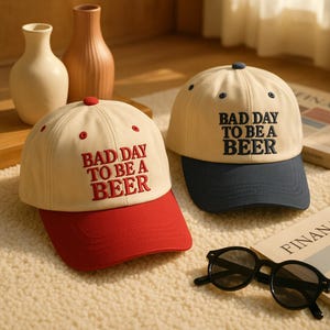Puede incluir: Dos gorras de béisbol con el texto "BAD DAY TO BE A BEER". Una gorra tiene una visera roja y la otra una visera azul marino. Ambas gorras son beige con el texto bordado en rojo y azul marino, respectivamente. Gafas de sol negras en primer plano.