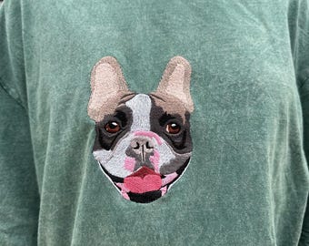 May include: A green t-shirt featuring an embroidered French Bulldog face. The dog's face has brown, black, white, and pink details. The shirt has a slightly faded appearance.