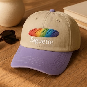 Puede incluir: Gorra de béisbol beige con visera y botón morados. Presenta un diseño de baguette arcoíris bordado y la palabra "faguette" en escritura blanca. Un accesorio casual.
