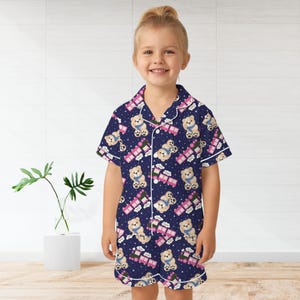 May include: Navy blue children's pyjama set featuring a pattern of teddy bears, pink trains, and white clouds. The short-sleeved top and shorts have white trim and buttons. The child is smiling.