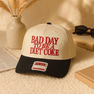 May include: Cream baseball cap with a black peak. The cap has the embroidered phrase "BAD DAY TO BE A DIET COKE" in red. A small label is visible on the peak. A fashionable accessory.