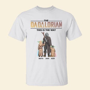 May include: White t-shirt featuring a graphic of a Mandalorian-inspired family. The design includes the text "THE DADALORIAN THIS IS THE WAY" above a depiction of a figure with two children, with the names Maya, John, and Alex below.