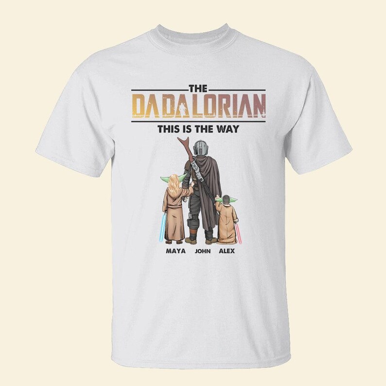May include: White t-shirt featuring a graphic of a Mandalorian-inspired family. The design includes the text "THE DADALORIAN THIS IS THE WAY" above a depiction of a figure with two children, with the names Maya, John, and Alex below.