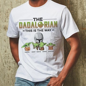 May include: White t-shirt featuring the text "THE DADALORIAN" with a graphic of Baby Yoda and the phrase "THIS IS THE WAY." Below the graphic are the names Laura, Kent, David, Anna, and Dustin. The shirt has a crew neck and short sleeves.