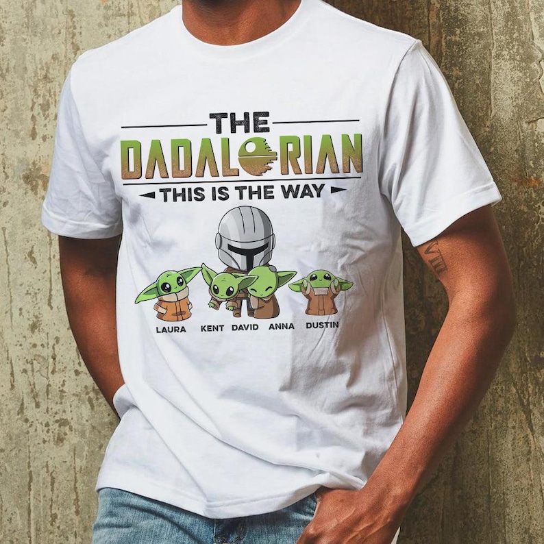 May include: White t-shirt featuring the text "THE DADALORIAN" with a graphic of Baby Yoda and the phrase "THIS IS THE WAY." Below the graphic are the names Laura, Kent, David, Anna, and Dustin. The shirt has a crew neck and short sleeves.