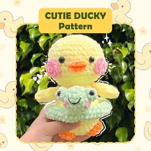 May include: A crocheted yellow duck with pink cheeks holding a green crocheted frog in a yellow and green crocheted inner tube. The duck is sitting on a hand and the background is green foliage. The text "CUTIE DUCKY Pattern" is at the top of the image.