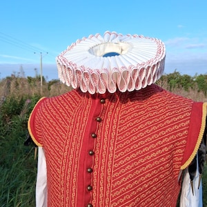 Könnte beinhalten: Eine rot-gold gemusterte Weste mit dunklen Knöpfen und einem weißen Rüschenkragen mit roter Bordüre. Das Kostüm ist vor dem Hintergrund eines blauen Himmels und eines grünen Feldes platziert, was auf historische Kleidung hindeutet.