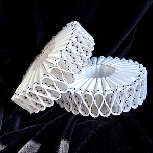 May include: Two white, ruffled Elizabethan collars with black decorative stitching. The collars are made of a pleated fabric, with a circular shape. The collars are displayed on a dark, textured surface.