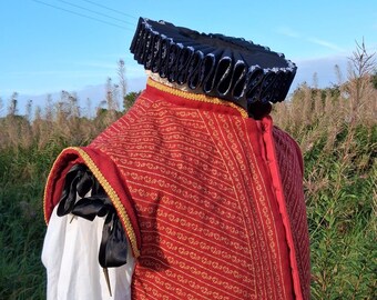 May include: A red and gold patterned vest with gold trim and black accents, paired with a black ruffled collar. The costume is set against a backdrop of tall grass and a blue sky, evoking a historical or theatrical theme.