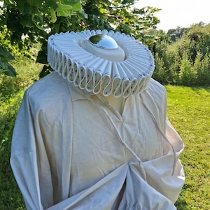 Könnte beinhalten: Ein cremefarbenes Hemd im Renaissance-Stil und ein gerüschter Kragen. Der Kragen ist weiß mit einem schwarzen, verzierten Design am Rand. Das Hemd hat einen V-Ausschnitt und lange Ärmel. Das Outfit wird auf einer Schaufensterpuppe präsentiert.