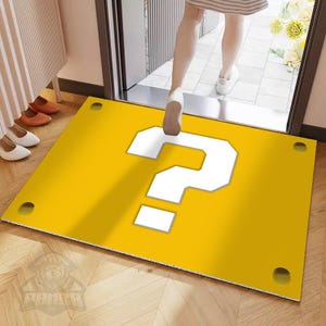 May include: A rectangular yellow floor mat featuring a large white question mark in the centre. The mat has four dark circular accents, one located near each corner. The design mimics a video game block, providing a flat surface for entryways.