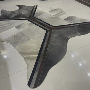 Puede incluir: Una alfombra de piel de vaca en tonos grises, blancos y negros, con un diseño de patchwork geométrico. La alfombra presenta un diseño central con rayas marrones y negras, añadiendo un toque moderno al material natural. Es una cubierta de suelo decorativa.