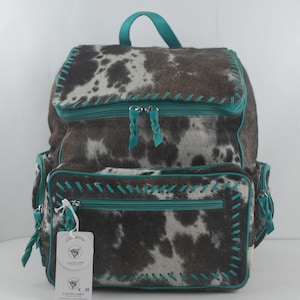 May include: A brown and white cowhide backpack with turquoise leather trim and stitching. The backpack has two large zippered compartments and a smaller zippered pocket on the front. The backpack has a tag that says "Cart Of Leather" and "# 30".